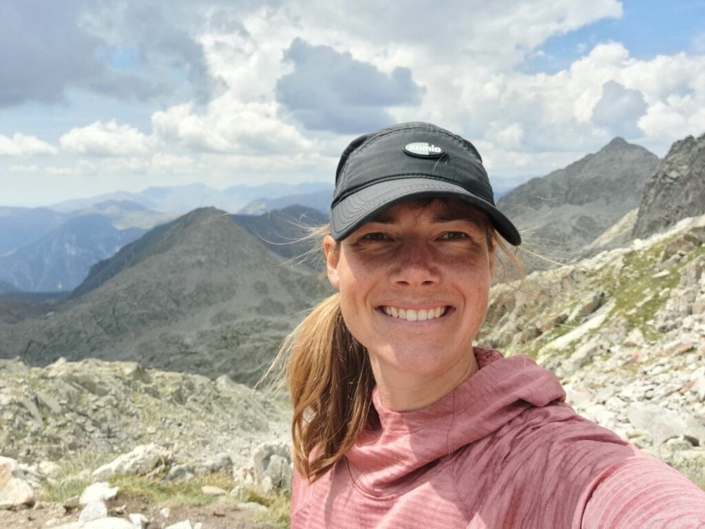 Selfie on top of a mountain pass with storm clouds and mountains in the background, wearing a black ballcap and a wide grin